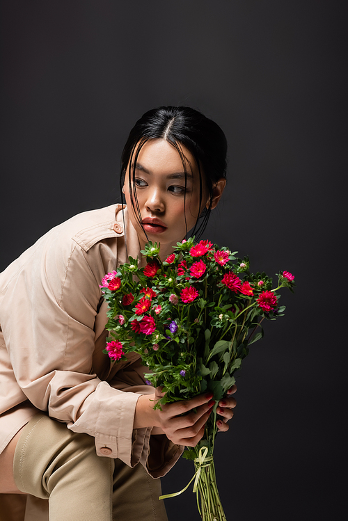 Trendy asian woman in boots and trench coat holding colorful flowers isolated on black