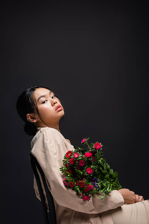 Stylish asian woman in trench coat and beige dress holding flowers while sitting on chair isolated on black