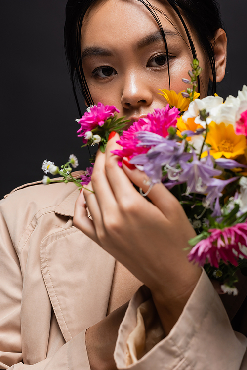 Stylish asian model in beige trench coat holding blurred flowers isolated on black