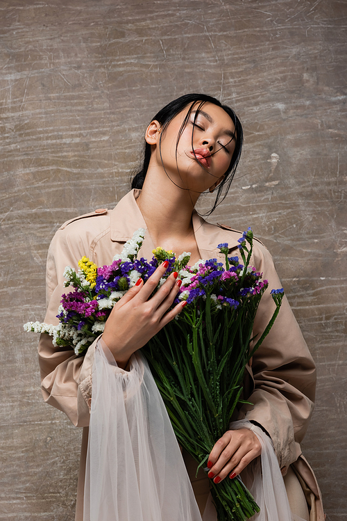 Low angle view of stylish asian woman in trench coat holding wildflowers on abstract brown background