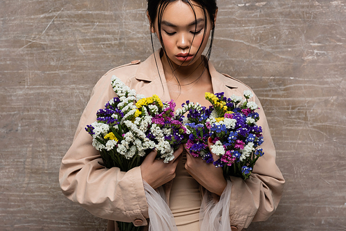 Young asian woman in beige trench coat holding wildflowers on abstract brown background