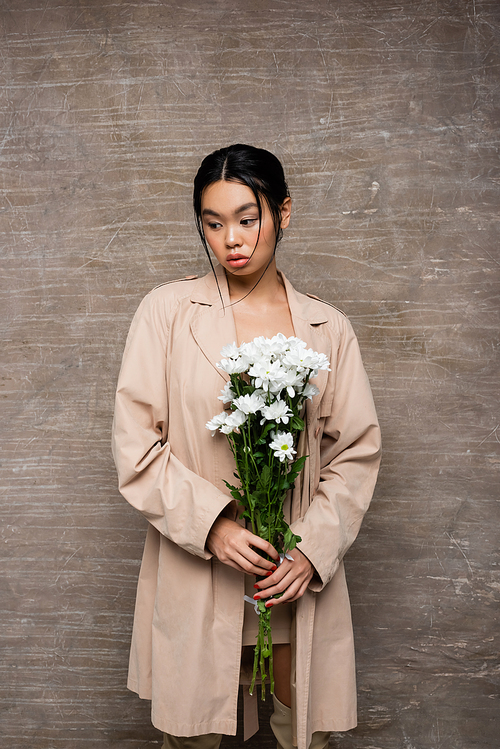 Asian woman in trench coat holding white chrysanthemums on abstract brown background