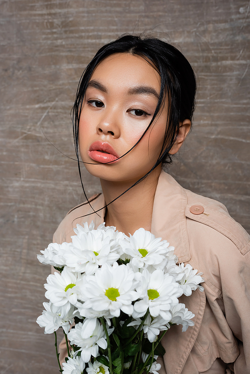 Pretty asian woman in trench coat holding white chrysanthemums on abstract brown background