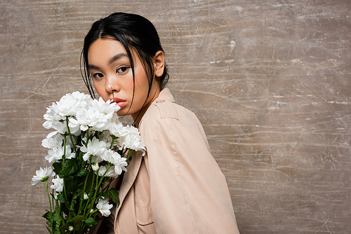 Stylish asian woman in trench coat looking at camera near beige chrysanthemums on abstract brown background