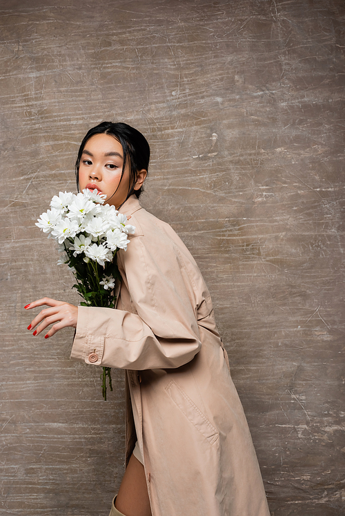 Pretty asian woman in trench coat posing with white chrysanthemums on abstract brown background