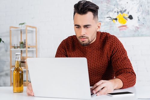 focused gambler looking at laptop near bottle of beer and smartphone