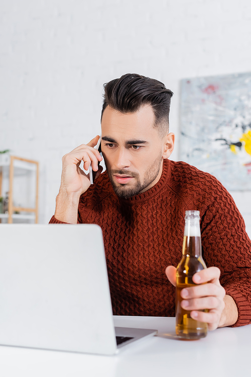 serious bookmaker with bottle of beer talking on smartphone near laptop