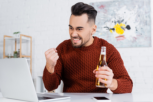 excited gambler with bottle of beer showing win gesture near laptop