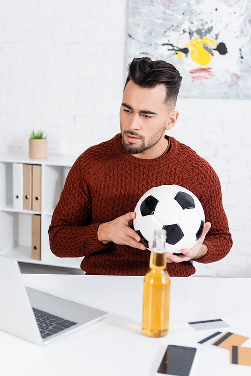 concentrated bookmaker with soccer ball looking at laptop near bottle of beer and credit cards