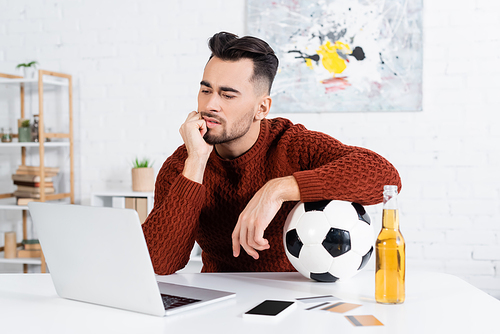 thoughtful bookmaker with soccer ball looking at laptop near smartphone and credit cards