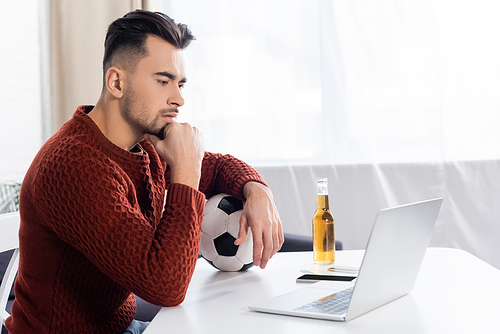 focused bookmaker with soccer ball looking at laptop near bottle of beer