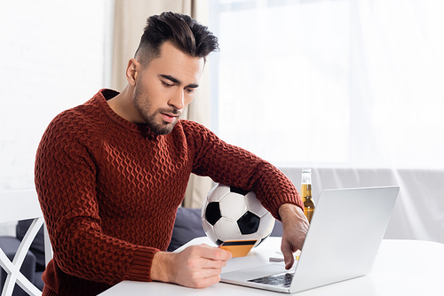 young bookmaker holding credit card near laptop and soccer ball