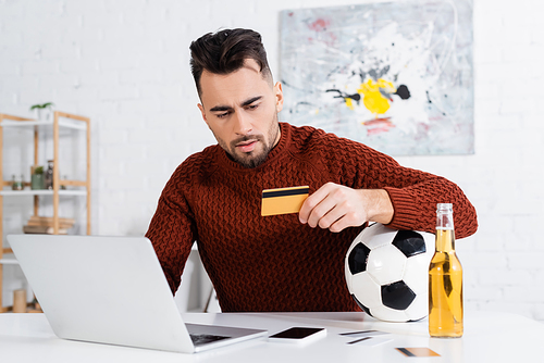 serious bookmaker holding credit card near soccer ball, laptop and beer