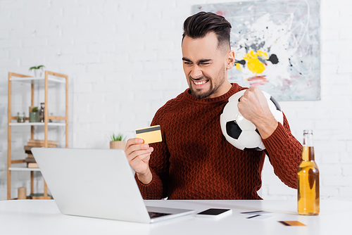 excited bookmaker with credit card and soccer ball near laptop and bottle of beer