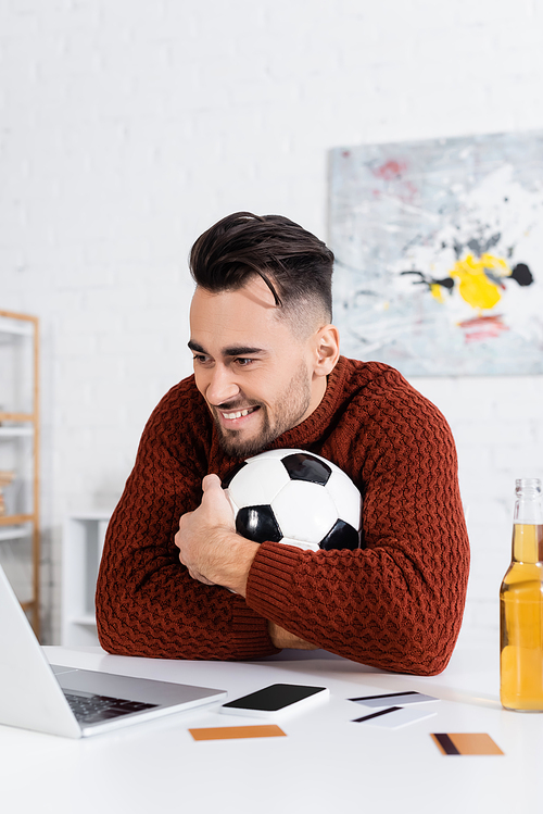 cheerful gambler hugging soccer ball near laptop, credit cards and beer