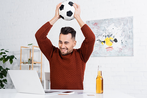 joyful bookmaker holding soccer ball above head near laptop and beer