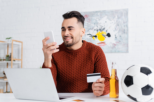 smiling gambler with credit card looking at smartphone near laptop, beer and soccer ball