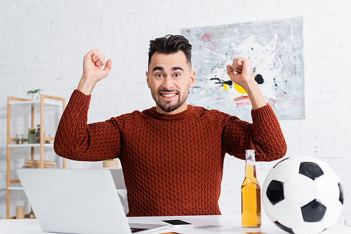 excited bookmaker showing success gesture near laptop, beer and soccer ball
