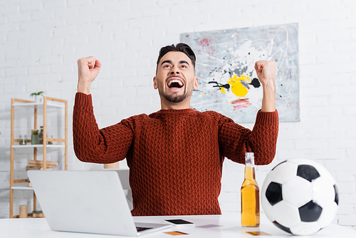 overjoyed gambler showing win gesture near laptop, soccer ball and beer