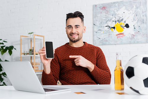 smiling gambler pointing at smartphone with blank screen near laptop, beer and blurred soccer ball