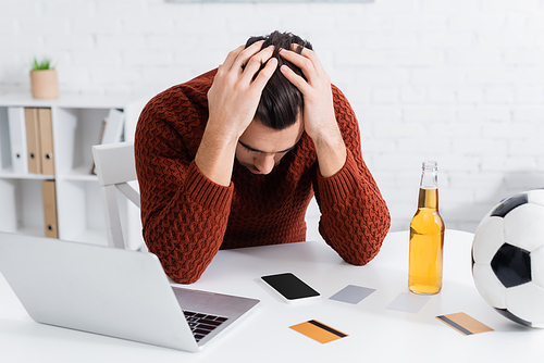 frustrated bookmaker sitting with bowed head near computer, credit cards and soccer ball