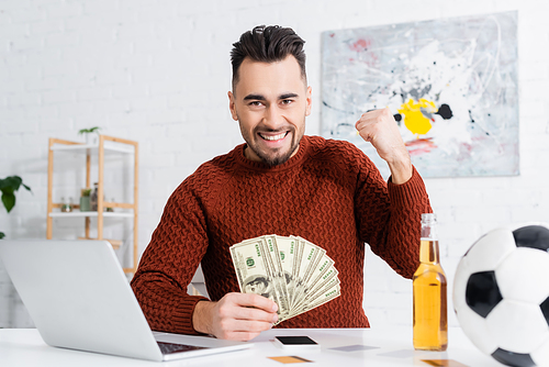 excited bookmaker with dollar banknotes showing win gesture near laptop and soccer ball