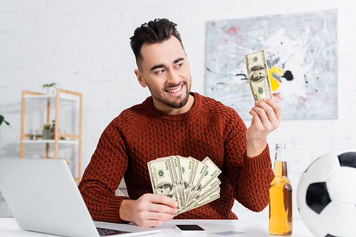 happy gambler with dollar banknotes near laptop, beer and soccer ball