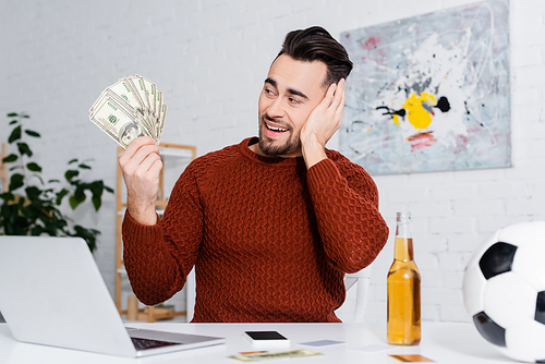 amazed bookmaker holding dollars near laptop, bottle of beer and soccer ball