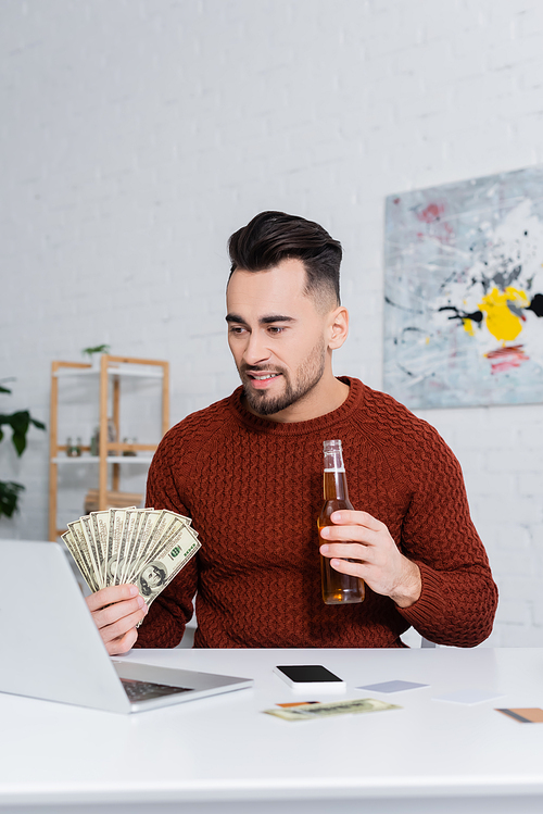 smiling gambler with dollars and bottle of beer near laptop and smartphone with blank screen