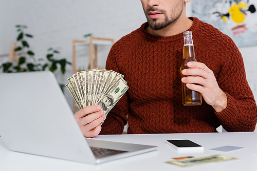 partial view of gambler holding dollars and beer near blurred laptop