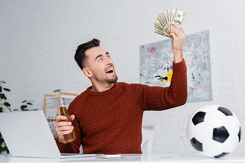 astonished gambler holding dollars and bottle of beer near laptop and soccer ball