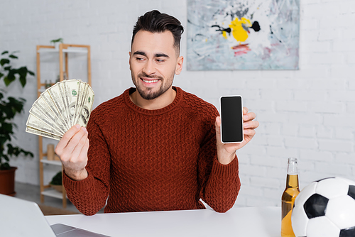 cheerful gambler holding dollars and smartphone with blank screen near soccer ball