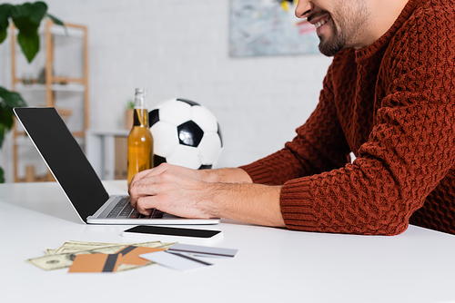 cropped view of smiling bookmaker typing on laptop near credit cards and soccer ball