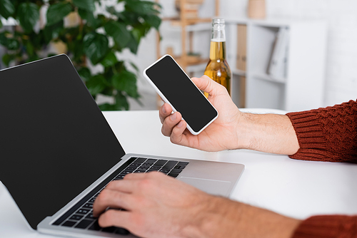 cropped view of gambler with smartphone typing on laptop with blank screen