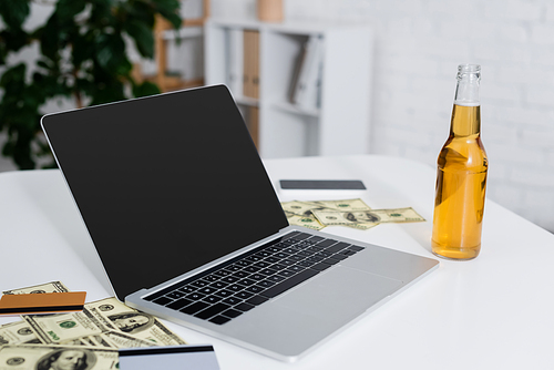 laptop with blank screen near credit cards, dollars and bottle of beer