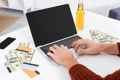 partial view of gambler typing on laptop with blank screen near dollars and credit cards