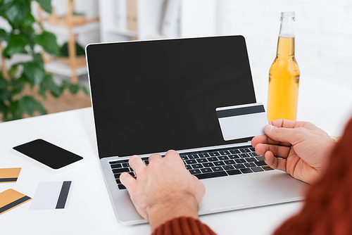 partial view of bookmaker near laptop with blank screen, credit cards and beer