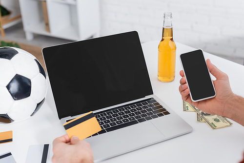 partial view of gambler with credit card and smartphone near laptop with blank screen