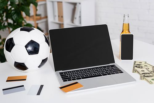 laptop with blank screen near soccer ball, credit cards, dollars and bottle of beer