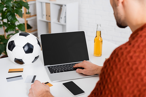 cropped view of bookmaker near laptop with blank screen, soccer ball and credit cards