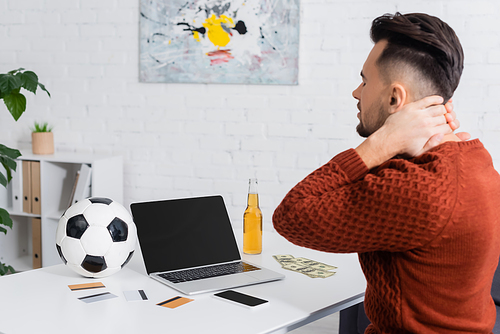 upset gambler sitting near laptop with blank screen, credit cards and soccer ball