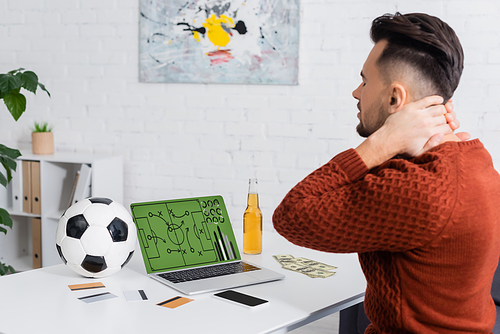 upset bookmaker touching neck near credit cards and laptop with sports game strategy on screen