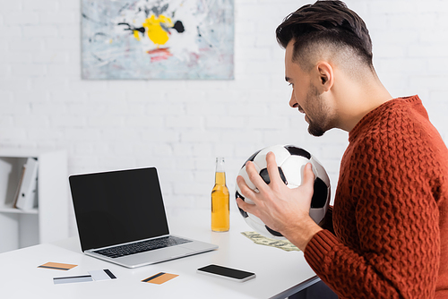 side view of gambler with soccer ball near laptop with blank screen, credit cards and bottle of beer
