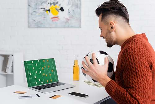 side view of bookmaker with soccer ball near credit cards and laptop with sports game strategy on screen