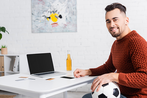 happy gambler with soccer ball looking at camera near laptop and credit cards