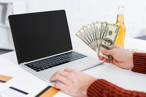 partial view of bookmaker with dollar banknotes near laptop with blank screen