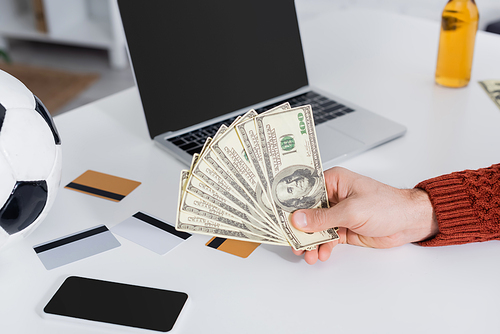 partial view of bookmaker holding dollars near credit cards, soccer ball and laptop