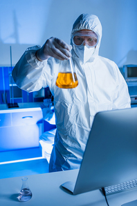scientist in hazmat suit holding flask with orange liquid near blurred computer monitor in lab