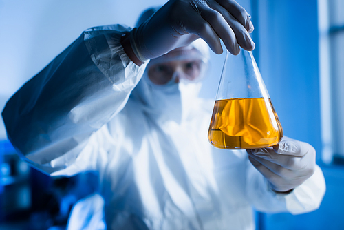 blurred biotechnologist in hazmat suit holding flask with orange liquid