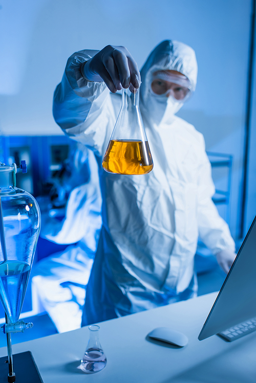 blurred scientist in hazmat suit holding flask with orange liquid in laboratory